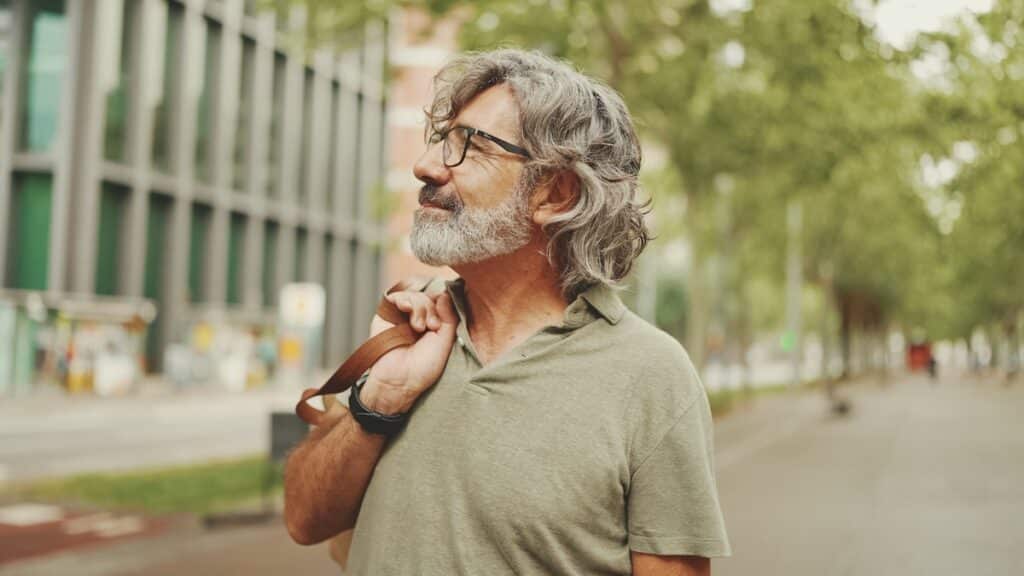 Middle aged man with gray hair and beard walks and looks around. Mature gentleman in eyeglasses with bag on his shoulder walks through the square on the cityscape background