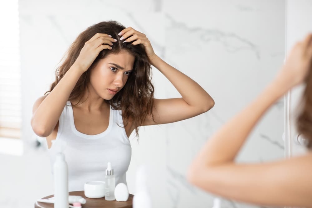 Frustrated young lady looking at her hair in the mirror and concerned with hair loss, in need LLLT.